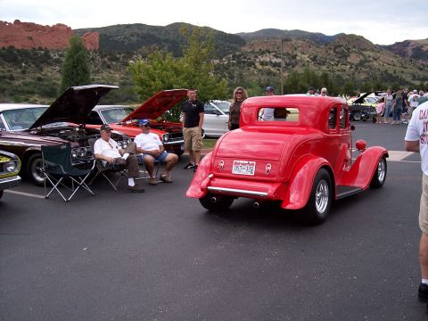 1972 Ford Ranchero - A red coupe hot rod is parked at a car show with other vehicles and people present.