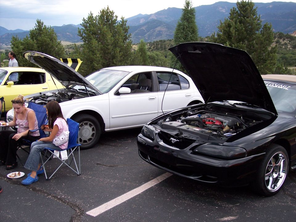 Black Ford Mustang, hood open, showing engine. White sedan and yellow car also visible