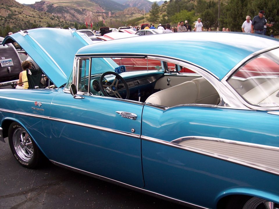 Blue 1957 Chevrolet Bel Air coupe with hood open, parked at an outdoor car show with mountains in background