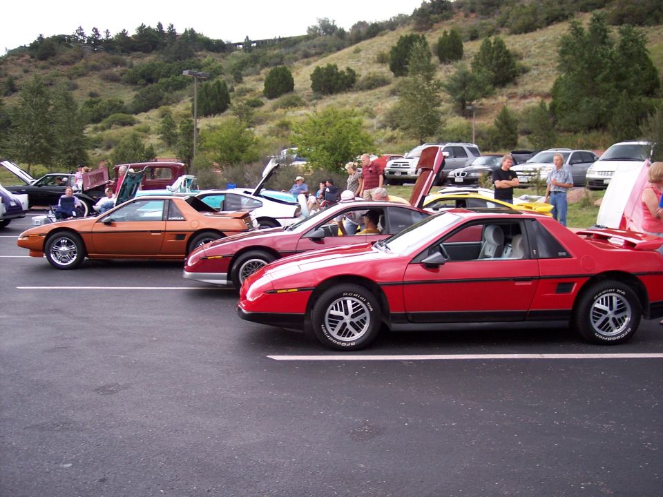Red Pontiac Fiero parked in a lot, side view, with gullwing doors open on other cars