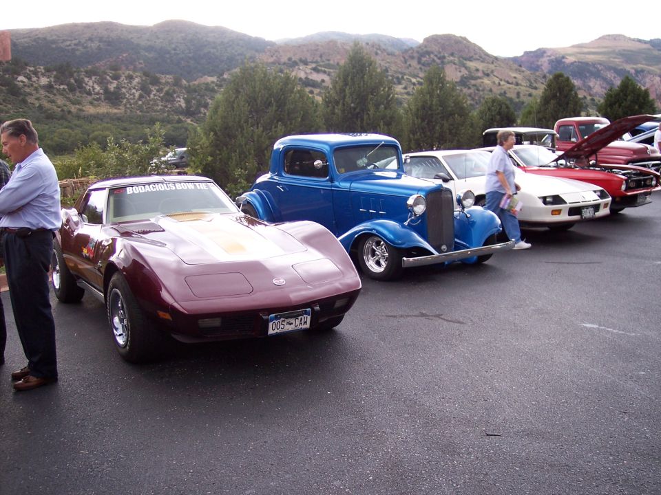 Maroon Corvette with 'BODACIOUS BOW TIE' on windshield, parked next to blue hot rod and other cars