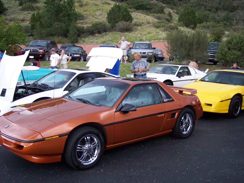 Orange Pontiac Fiero parked next to a yellow Fiero and a white car with hood up