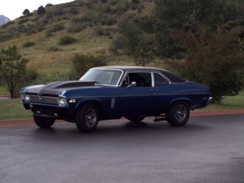 1972 Ford Ranchero - A blue 1970 Chevrolet Nova coupe with a black hood scoop and racing stripes is parked on asphalt with a hilly, tree-covered background.