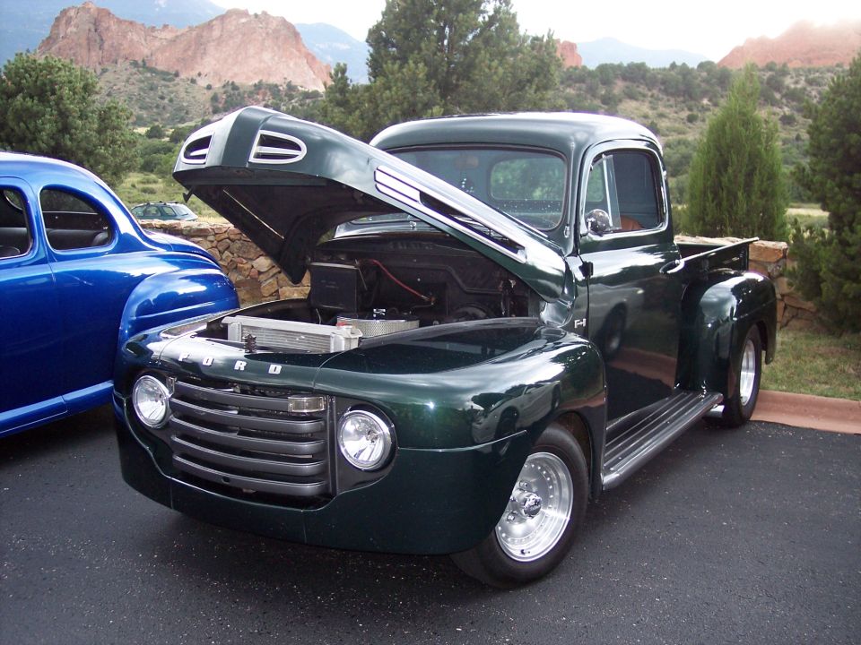 Green Ford F-series truck with hood open, parked next to a blue car. Mountains in background