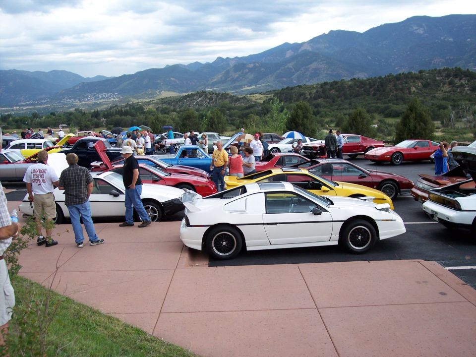 White sports car with a large rear spoiler parked in a lot with other cars and mountains in the background