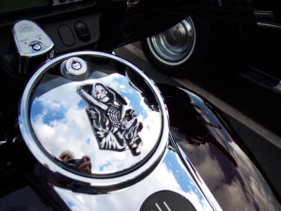 Motorcycle gas tank with chrome cover showing grim reaper emblem. Reflection shows sky and person with camera