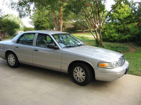 2003 Ford Crown Victoria - A silver 2003 Ford Crown Victoria sedan is parked outdoors on a concrete driveway with trees and foliage in the background.