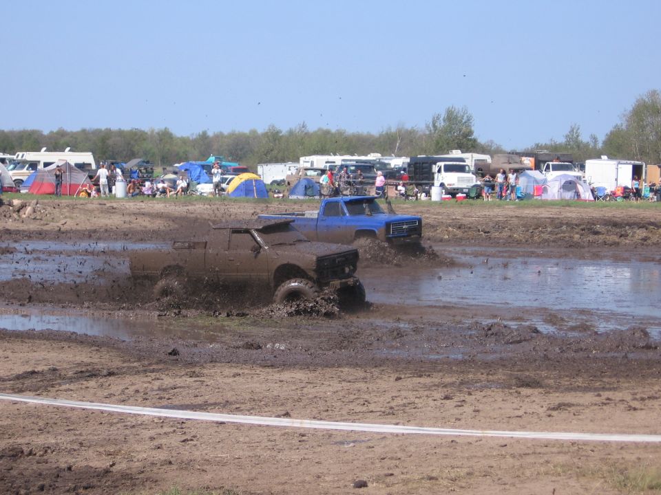 Muddy brown pickup truck and blue pickup truck driving through mud pit with spectators and tents in background