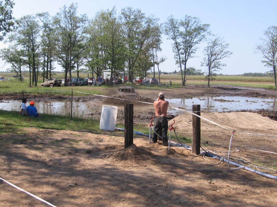 Mud bogging event with people watching and a vehicle in the mud pit. A man stands near water spigots