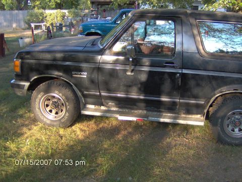1987 Ford Bronco - A black Ford Bronco is parked in a yard, with a blue SUV visible in the background.