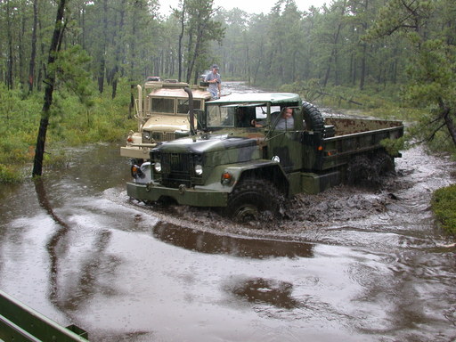 Two military trucks driving through a muddy, flooded wooded area. One truck is green, the other is tan