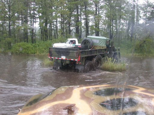 Military truck with a white pickup truck in its bed, driving through water