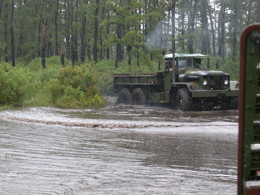 Military truck with multiple axles driving through a flooded wooded area. Another vehicle is partially visible on the right