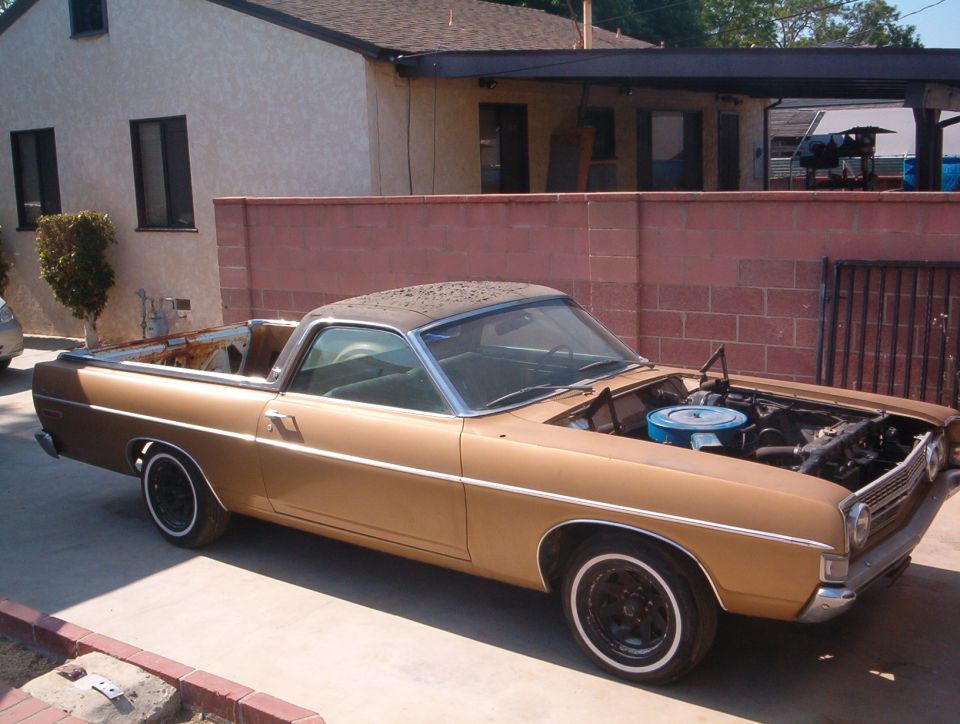 Gold 1968 Ford Ranchero with hood removed showing engine, parked by a brick wall