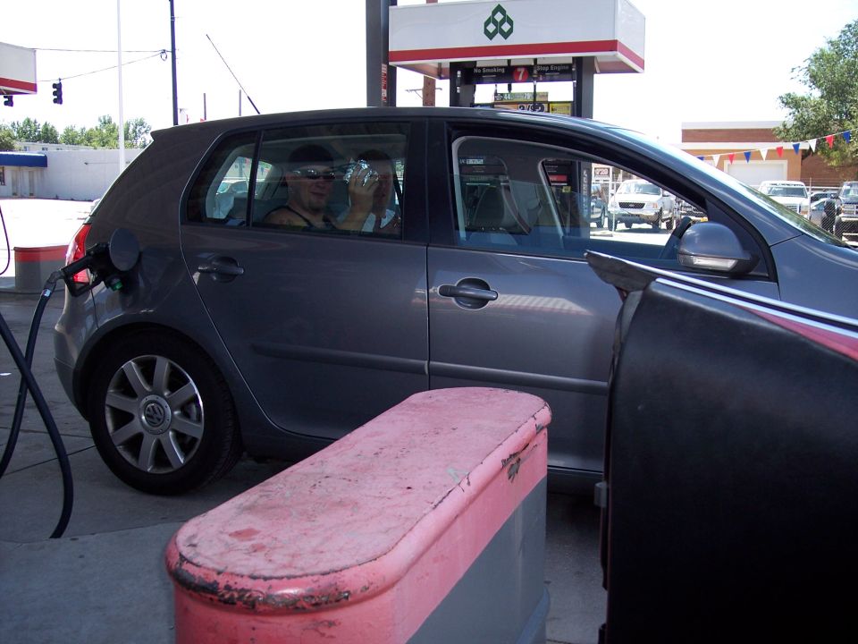Gray Volkswagen Golf with gas pump nozzle in fuel door, two people inside, at a gas station