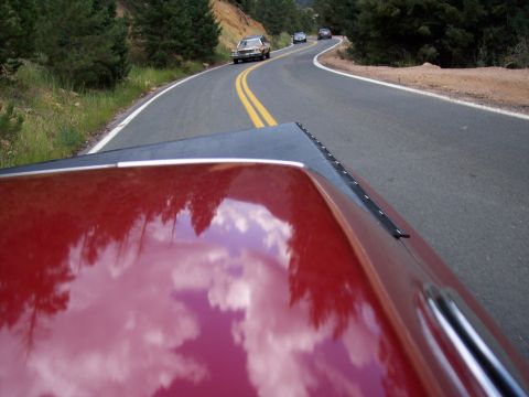 1972 Ford Ranchero - A red vehicle's hood reflects the sky and surrounding trees as it travels on a winding mountain road behind other cars.