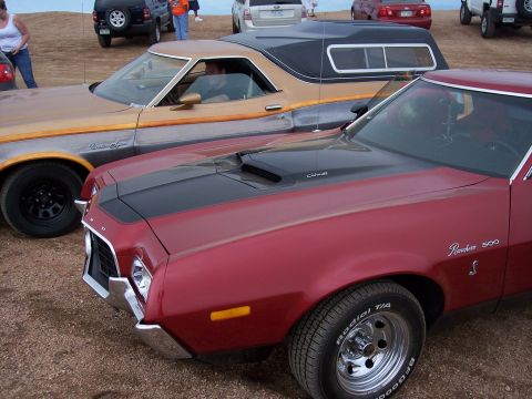 1972 Ford Ranchero - Two Ford Rancheros are parked side-by-side on a dirt lot with other vehicles and people in the background.