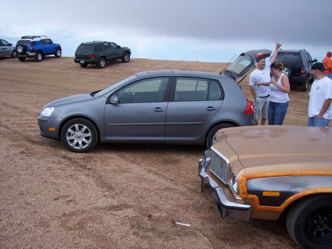 1972 Ford Ranchero - Multiple vehicles parked on a dirt lot with people standing around the grey hatchback.