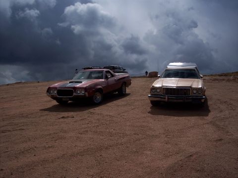 1972 Ford Ranchero - Two vehicles, a red Ranchero and a beige station wagon, sit parked on a dirt surface with dramatic clouds overhead.