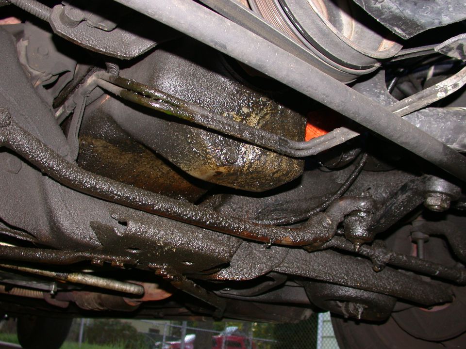 Underside of a car showing extensive oil leaks around the engine and suspension components
