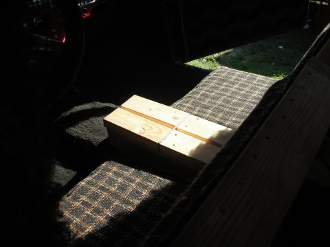 1998 Ford Crown Victoria - Pine wooden blocks and boards staged on a checkered carpet mat inside a dim vehicle interior, with sunlit grass visible beyond an open door.