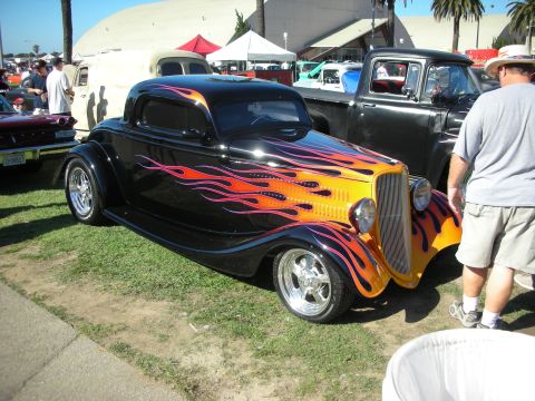 1998 Ford Crown Victoria - black three-window hot rod coupe with orange-and-purple flame paint and chrome wheels parked on grass at a car show.