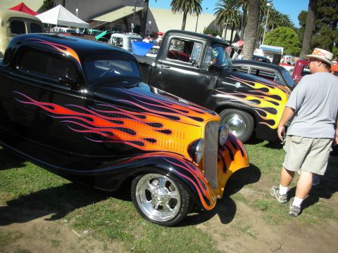 1998 Ford Crown Victoria - Black hot rod coupe with vivid flame paint and chrome wheel, parked next to a similarly flamed pickup on grass as a spectator walks by.