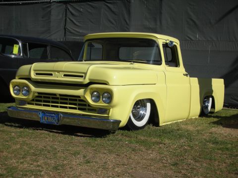 1998 Ford Crown Victoria - lowered yellow Ford F-100 stepside pickup with quad headlights and chrome wheels on whitewall tires, sitting on grass near a tent backdrop.