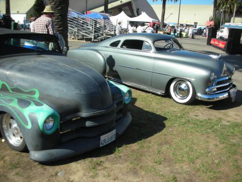 1998 Ford Crown Victoria - primered custom with green flames parked nose-to-nose with a silver chopped early-1950s lead sled on whitewalls at an outdoor show.