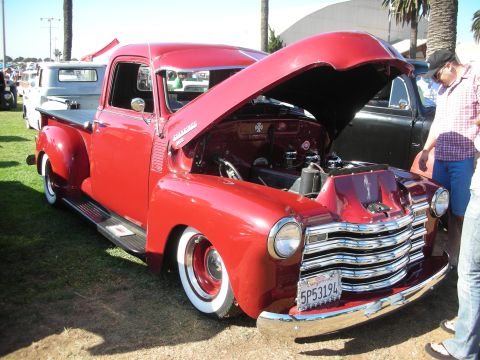 1998 Ford Crown Victoria - glossy red early-1950s Chevrolet pickup, hood raised to show its engine, lowered on whitewalls with a spectator looking on.