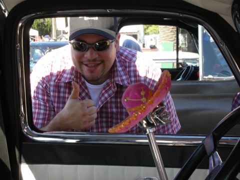 1998 Ford Crown Victoria - smiling man in a plaid shirt, cap and sunglasses gives a thumbs-up through the open window of a custom car with a pink sombrero shifter.