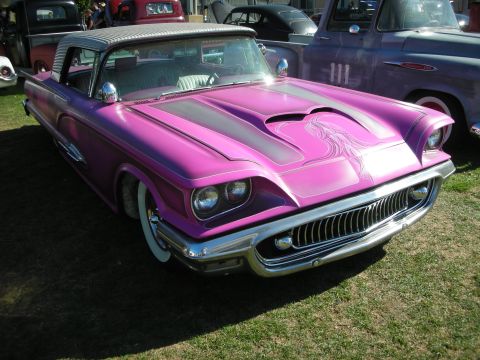 1998 Ford Crown Victoria - pink-and-purple custom Thunderbird with airbrushed hood scoop, chrome grille and whitewall tires displayed among other customs on grass.
