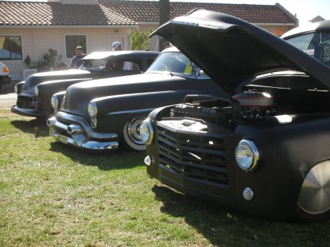 1998 Ford Crown Victoria - lineup of matte-black chopped customs on grass, with a black Studebaker-style pickup, hood raised, in the foreground by a stucco building.