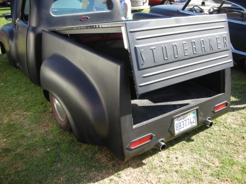 1998 Ford Crown Victoria - Open tailgate of a satin-black Studebaker pickup, lettered STUDEBAKER, revealing a carpeted bed and dual exhaust tips at a car show.