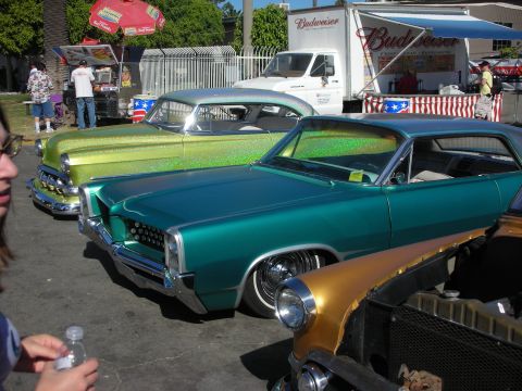 1998 Ford Crown Victoria - lowered teal-green custom coupe sits beside other lowriders at an outdoor car show, with a Budweiser box truck and tents in the background.