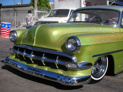 1998 Ford Crown Victoria - Close-up of the chrome grille, bullet headlight and green metalflake fender of a lowered 1950s custom car on a sunny lot.