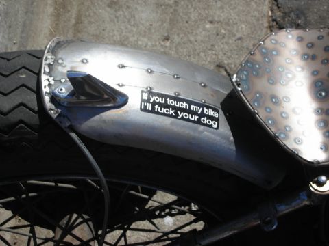 1998 Ford Crown Victoria - Close-up of a riveted bare-metal motorcycle rear fender bearing a black sticker that reads 'If you touch my bike I'll fuck your dog.'