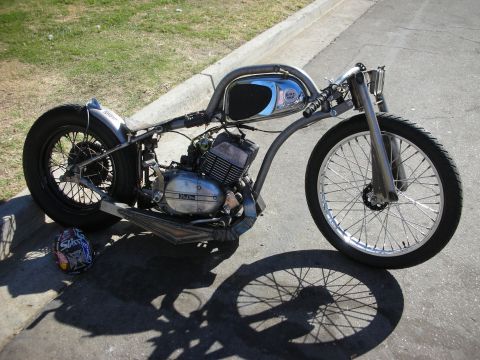 1998 Ford Crown Victoria - long bare-metal rigid chopper with a single-cylinder engine and chrome tank rests on pavement beside a graphic helmet.