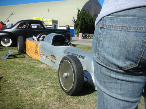 1998 Ford Crown Victoria - bare-aluminum streamliner lakester lettered 'Vintage Hammer Garage' sits on a show field as a person in jeans stands in the foreground.
