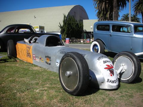 1998 Ford Crown Victoria - polished bare-aluminum streamliner racer marked 'Bean Bandits' sits on grass flanked by vintage hot rods at an outdoor show.