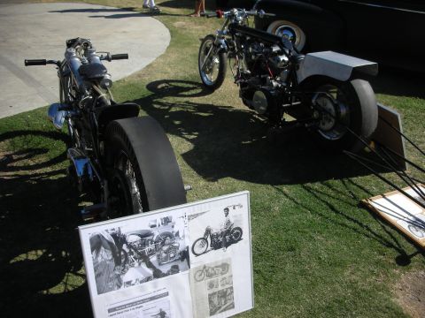 1998 Ford Crown Victoria - Two long custom dragster-style motorcycles sit on grass beside a printed black-and-white info display board at a show.