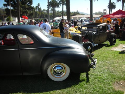 1998 Ford Crown Victoria - matte-black chopped coupe on wide whitewalls sits among hot rods at a busy outdoor car show under palm trees.