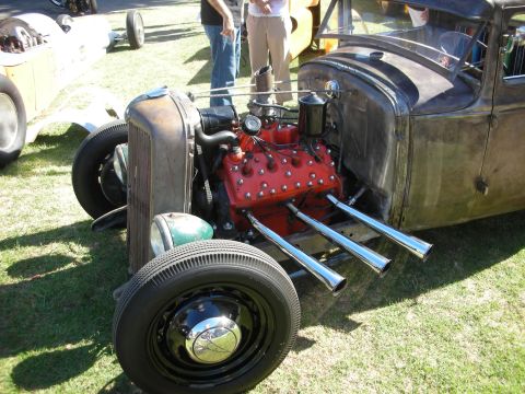 1998 Ford Crown Victoria - rusty patina rat rod displays a red-painted flathead V8 with chrome zoomie exhaust pipes on a grass show field.