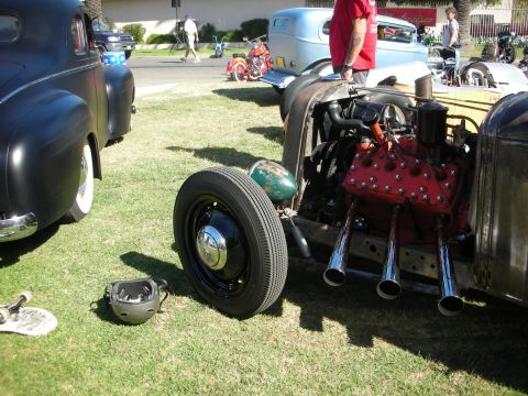 1998 Ford Crown Victoria - patina rat rod with an exposed red flathead V8 and chrome zoomie pipes sits on grass with a helmet beside its bare wheel.