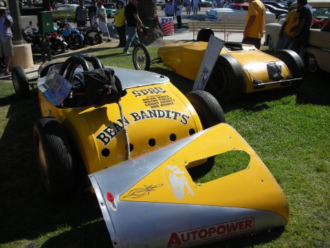 1998 Ford Crown Victoria - Two yellow 'Bean Bandits' streamliner land-speed racers sit on grass with an Autopower sticker as a crowd mills behind them.