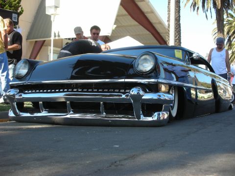 1998 Ford Crown Victoria - slammed glossy black 1950s custom car sits nose-low at an outdoor show with onlookers and palm trees behind it.