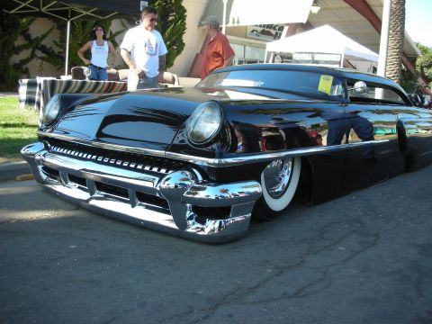 1998 Ford Crown Victoria - glossy black chopped and lowered 1950s custom coupe sits on whitewall tires under a show canopy as spectators look on.