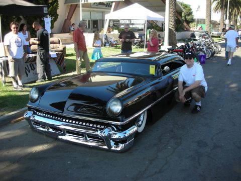 1998 Ford Crown Victoria - man crouches next to a low glossy black chopped custom coupe parked on pavement at a sunny outdoor car show.