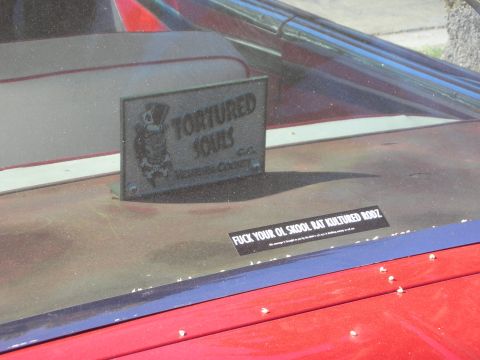 1998 Ford Crown Victoria - cast Tortured Souls Ventura County car-club plaque sits on a red car cowl beside a printed slogan strip.