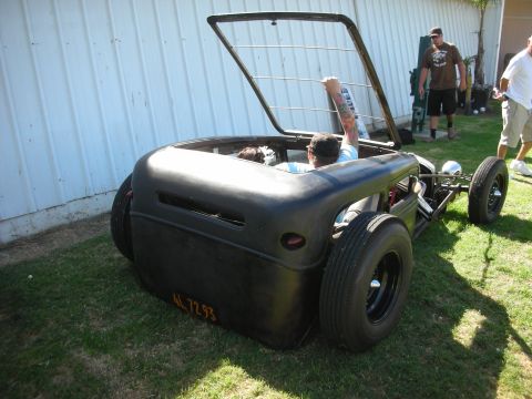 1998 Ford Crown Victoria - low flat-black channeled rat rod coupe with its trunk lid raised sits on wide tires on grass beside a metal shed.